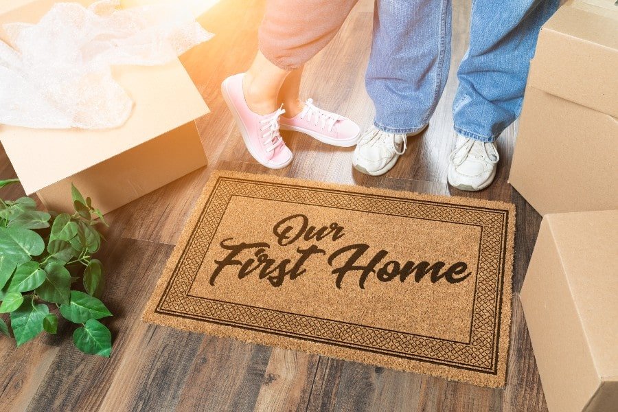 Couple standing behind a welcome mat that says Our First Home